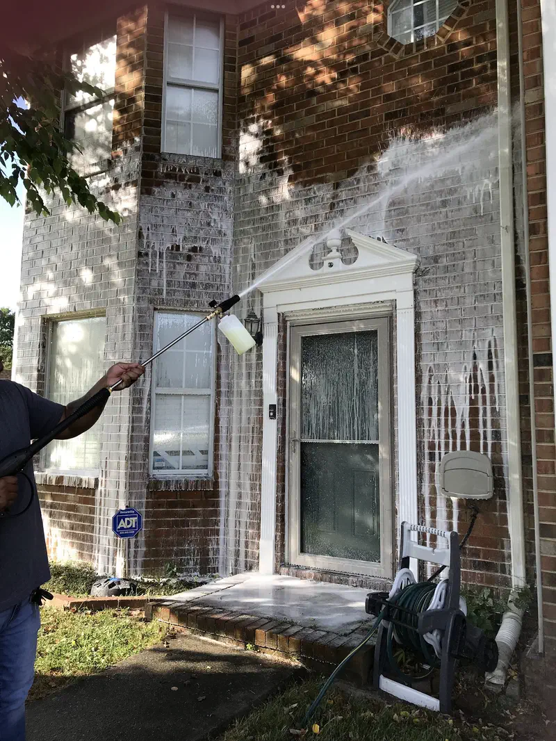 A brick house front covered in cleaning foam, the wand spraying the wall